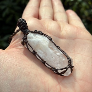 Puede incluir: Un colgante de cristal claro y alargado envuelto en macramé marrón oscuro. El cristal es de un color pálido y translúcido. El colgante está suspendido de un cordón marrón a juego. El fondo es un follaje verde borroso.