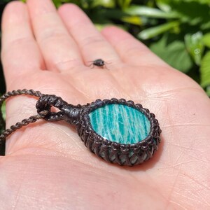 May include: A handcrafted macrame pendant necklace featuring a round, polished amazonite stone with a teal and white striped pattern. The stone is set in a dark brown macrame weave, suspended from a matching cord.