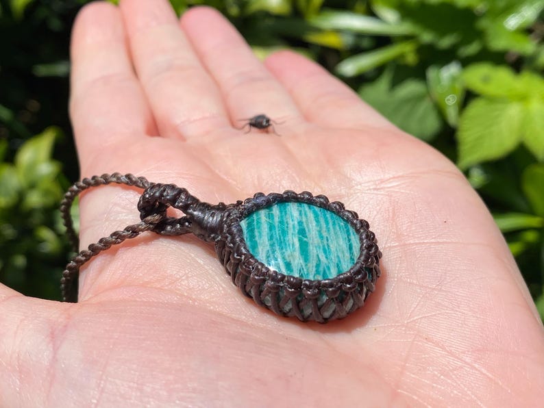 May include: A handcrafted macrame pendant necklace featuring a round, polished amazonite stone with a teal and white striped pattern. The stone is set in a dark brown macrame weave, suspended from a matching cord.
