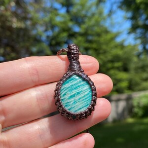 May include: A handcrafted pendant featuring a polished, oval-shaped amazonite stone with a light blue and white striped pattern. The stone is encased in a dark brown macrame setting, creating a unique, earthy aesthetic. The pendant is suspended from a matching macrame cord.