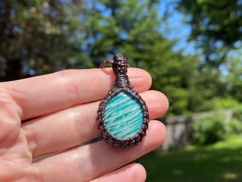 May include: A handcrafted pendant featuring a polished, oval-shaped amazonite stone with a light blue and white striped pattern. The stone is encased in a dark brown macrame setting, creating a unique, earthy aesthetic. The pendant is suspended from a matching macrame cord.