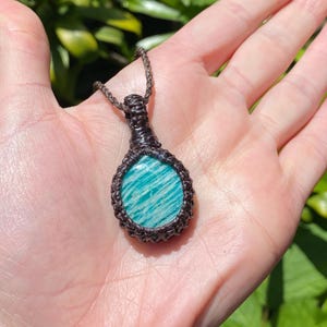 May include: A handcrafted pendant necklace featuring a polished, oval-shaped amazonite stone with blue-green stripes. The stone is encased in a dark brown macrame setting and suspended from a matching cord. The necklace is displayed against a natural background.