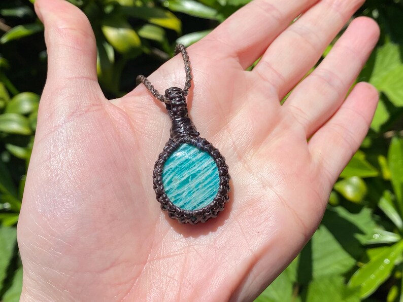May include: A handcrafted pendant necklace featuring a polished, oval-shaped amazonite stone with blue-green stripes. The stone is encased in a dark brown macrame setting and suspended from a matching cord. The necklace is displayed against a natural background.