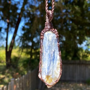 May include: A blue kyanite crystal pendant wrapped in copper wire, suspended from a brown cord. The crystal has a layered appearance with streaks of white and yellow. The background is blurred, with green foliage and sunlight.