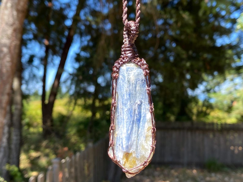 May include: A blue kyanite crystal pendant wrapped in copper wire, suspended from a brown cord. The crystal has a layered appearance with streaks of white and yellow. The background is blurred, with green foliage and sunlight.