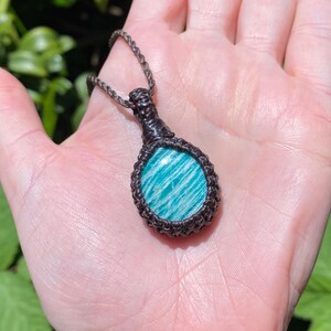 May include: A handmade macrame necklace featuring a round, polished amazonite stone with a blue and white striped pattern. The stone is encased in a dark brown macrame setting and hangs from a matching cord. The necklace is displayed against a natural green background.