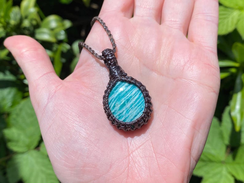 May include: A handmade macrame necklace featuring a round, polished amazonite stone with a blue and white striped pattern. The stone is encased in a dark brown macrame setting and hangs from a matching cord. The necklace is displayed against a natural green background.