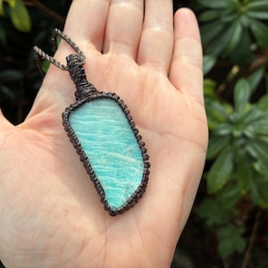 Pode incluir: Pingente retangular de pedra amazonita azul clara com um cord&atilde;o de macram&ecirc; marrom. A pedra tem um padr&atilde;o &uacute;nico de linhas azuis claras e escuras. O pingente &eacute; segurado em uma m&atilde;o contra um fundo de folhagem verde.