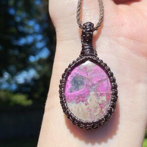 May include: A handcrafted pendant necklace featuring a teardrop-shaped stone with pink and gray hues. The stone is encased in a dark brown macrame setting and suspended from a matching cord. The necklace is displayed against a blurred background.