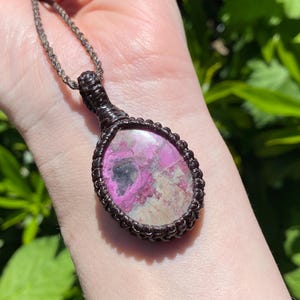 May include: A pendant necklace featuring a large, oval-shaped gemstone with pink and white hues. The stone is encased in a dark brown, woven macrame setting and hangs from a matching chain. The necklace is displayed against a blurred green background.