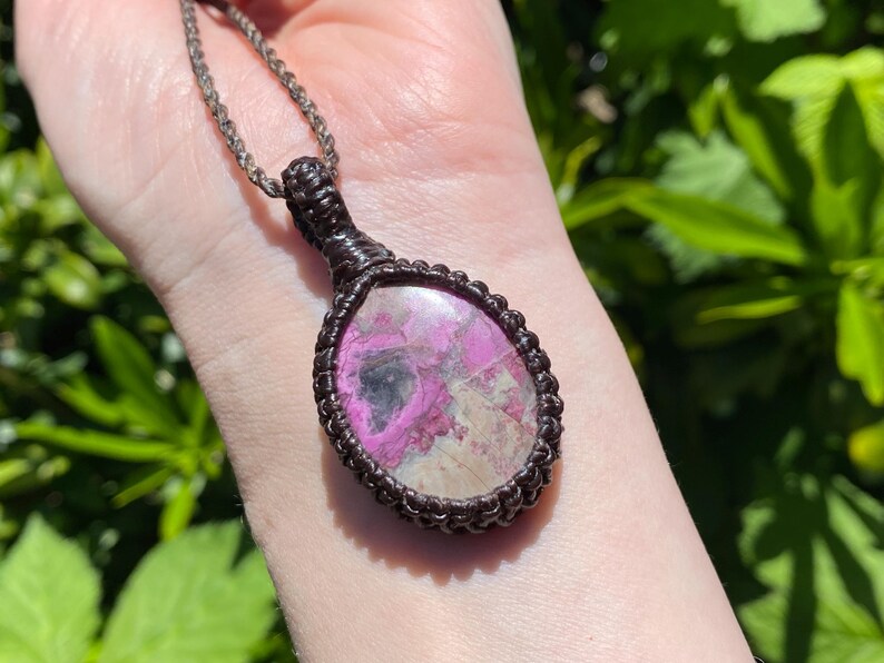 May include: A pendant necklace featuring a large, oval-shaped gemstone with pink and white hues. The stone is encased in a dark brown, woven macrame setting and hangs from a matching chain. The necklace is displayed against a blurred green background.
