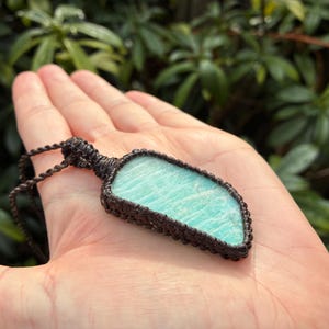 Pode incluir: Um colar de macram&eacute; artesanal com uma pedra de amazonita azul clara. A pedra &eacute; envolvida em uma trama de macram&eacute; marrom escuro e suspensa por um cord&atilde;o correspondente. O colar &eacute; exibido em um fundo verde desfocado.