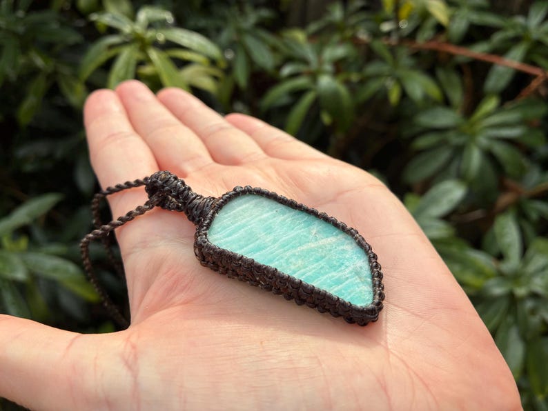 Pode incluir: Um colar de macram&eacute; artesanal com uma pedra de amazonita azul clara. A pedra &eacute; envolvida em uma trama de macram&eacute; marrom escuro e suspensa por um cord&atilde;o correspondente. O colar &eacute; exibido em um fundo verde desfocado.