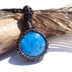 May include: A handcrafted pendant necklace featuring a round, bright blue gemstone. The stone is encased in a dark brown, woven macrame setting and suspended from a matching cord. The necklace is displayed on a piece of driftwood, with a feather in the background.