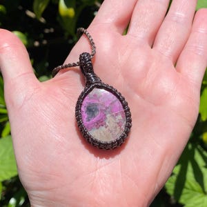 May include: A handcrafted pendant necklace featuring a large, oval-shaped stone with pink and gray hues, encased in a dark brown woven macrame setting. The pendant hangs from a matching woven cord, showcasing a natural, earthy aesthetic.