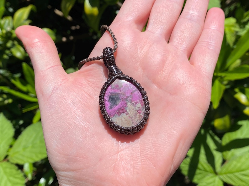 May include: A handcrafted pendant necklace featuring a large, oval-shaped stone with pink and gray hues, encased in a dark brown woven macrame setting. The pendant hangs from a matching woven cord, showcasing a natural, earthy aesthetic.