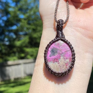 May include: A handcrafted pendant necklace featuring a pink and gray stone set in a brown macrame design. The pendant is oval-shaped and hangs from a matching brown cord. The background is a blurred view of green foliage.