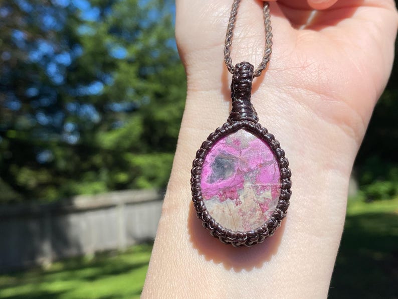 May include: A handcrafted pendant necklace featuring a pink and gray stone set in a brown macrame design. The pendant is oval-shaped and hangs from a matching brown cord. The background is a blurred view of green foliage.