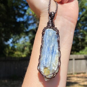 May include: A blue kyanite crystal pendant, wrapped in brown wire, hangs from a brown cord. The crystal has streaks of blue and white. The background is a blurred view of green trees and a wooden fence.