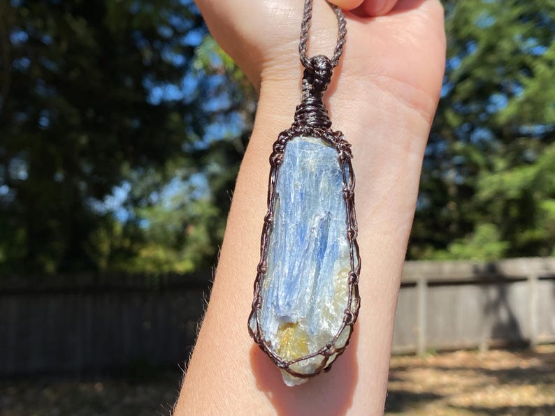 May include: A blue kyanite crystal pendant, wrapped in brown wire, hangs from a brown cord. The crystal has streaks of blue and white. The background is a blurred view of green trees and a wooden fence.