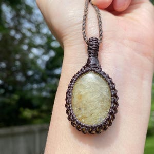 May include: A handcrafted macrame pendant necklace featuring a light-colored oval stone. The stone is encased in a dark brown macrame weave, suspended from a matching cord. The necklace is displayed against a blurred background.
