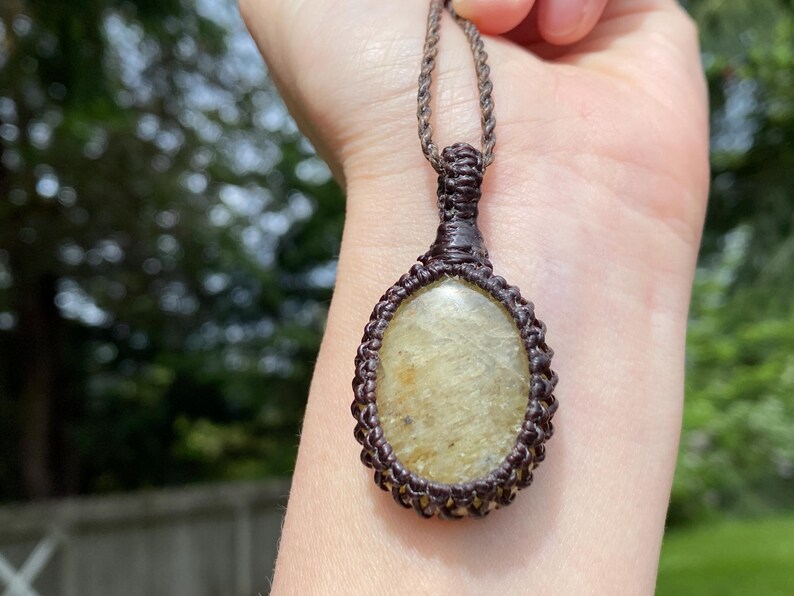 May include: A handcrafted macrame pendant necklace featuring a light-colored oval stone. The stone is encased in a dark brown macrame weave, suspended from a matching cord. The necklace is displayed against a blurred background.