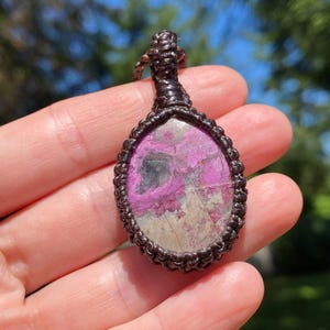 May include: A handcrafted pendant featuring a polished, oval-shaped stone with a vibrant pink and gray pattern. The stone is encased in a dark brown, intricately woven macrame setting, creating a unique and earthy aesthetic. The pendant is suspended from a matching cord.
