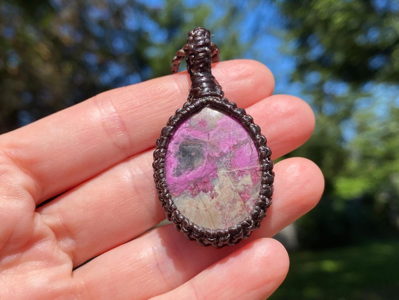 May include: A handcrafted pendant featuring a polished, oval-shaped stone with a vibrant pink and gray pattern. The stone is encased in a dark brown, intricately woven macrame setting, creating a unique and earthy aesthetic. The pendant is suspended from a matching cord.