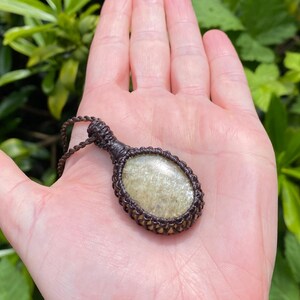 May include: A handcrafted pendant necklace featuring a light green, oval-shaped stone encased in a brown macrame setting. The necklace is displayed against a background of green foliage.
