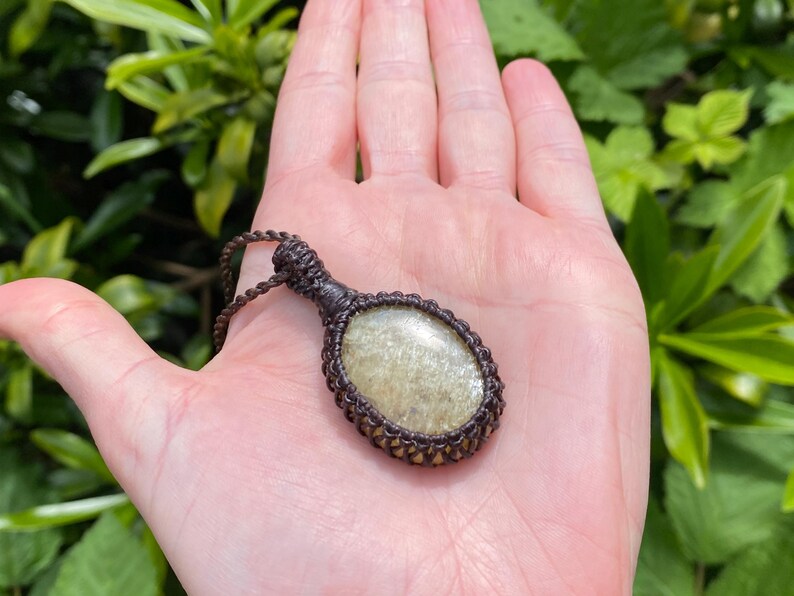 May include: A handcrafted pendant necklace featuring a light green, oval-shaped stone encased in a brown macrame setting. The necklace is displayed against a background of green foliage.