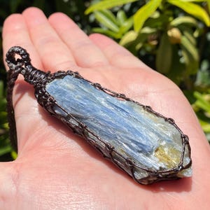 May include: A blue kyanite crystal pendant, wrapped in a dark brown woven macrame. The crystal has a layered appearance with shades of blue and a touch of gold. The pendant is suspended from a matching brown cord.