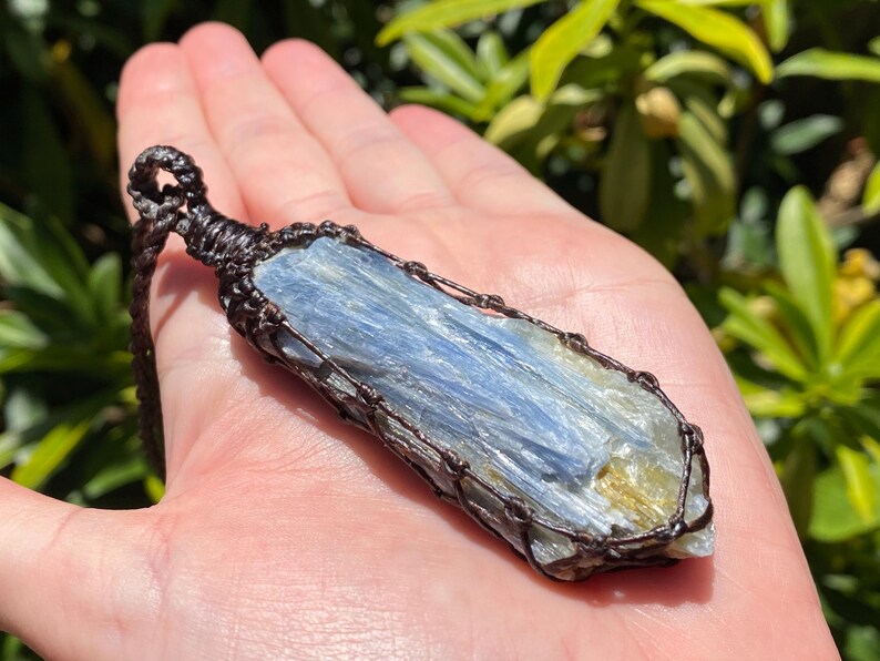 May include: A blue kyanite crystal pendant, wrapped in a dark brown woven macrame. The crystal has a layered appearance with shades of blue and a touch of gold. The pendant is suspended from a matching brown cord.
