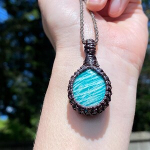 May include: A handcrafted pendant necklace featuring a teardrop-shaped amazonite stone. The stone is a vibrant turquoise color with white striations, encased in a dark brown macrame setting, suspended from a silver-toned chain.