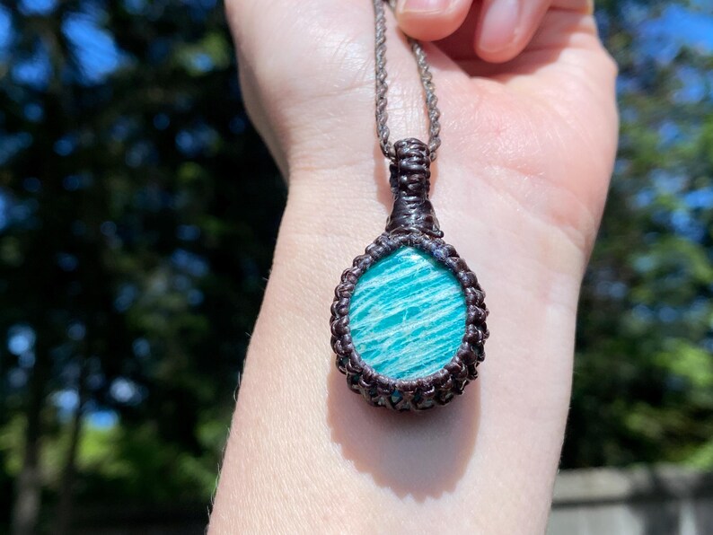 May include: A handcrafted pendant necklace featuring a teardrop-shaped amazonite stone. The stone is a vibrant turquoise color with white striations, encased in a dark brown macrame setting, suspended from a silver-toned chain.