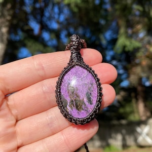 Puede incluir: Un colgante de piedra preciosa púrpura y verde con un cordón marrón. El colgante tiene forma ovalada y una textura natural y rugosa.