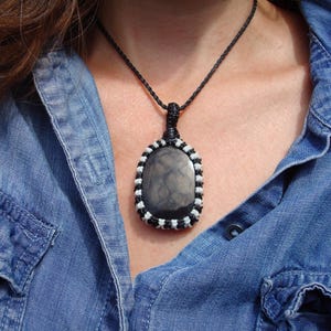 K&ouml;nnte beinhalten: Eine handgefertigte Halskette mit einem gro&szlig;en, ovalen Anh&auml;nger aus dunklem, marmoriertem Stein. Der Stein ist in einem Makramee-Design mit abwechselnd schwarzen und wei&szlig;en F&auml;den gefasst. Die Halskette hat eine schwarze Kordel.