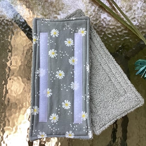 May include: A rectangular gray fabric pad featuring a white daisy floral pattern and two vertical strips of white hook-and-loop fastener. The reverse side of the pad is made of a textured gray terry cloth material with stitched edges.
