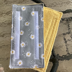 May include: A rectangular cleaning pad featuring a gray fabric top with a white and yellow daisy floral pattern and two vertical white hook-and-loop fastener strips. The underside of the pad is made of a textured yellow absorbent fabric material.