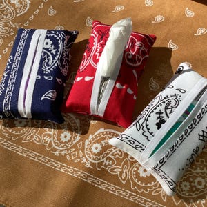May include: Three rectangular tissue box covers in red, white, and blue bandana patterns. Each cover has a white band across the front. The covers are on a brown paisley patterned surface.
