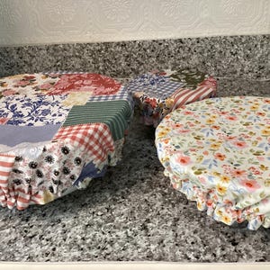 May include: Three round fabric bowl covers with elastic edges. The covers feature patchwork patterns with floral, gingham, and striped designs in red, blue, green, and white. The covers are on a granite countertop.