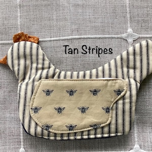 May include: A fabric chicken-shaped ornament featuring tan stripes and a tan wing patch with a repeating black bee pattern. The chicken has a small orange patterned comb and beak. The text Tan Stripes is printed above the chicken figure.