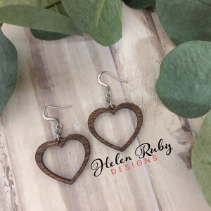 May include: A pair of wooden heart-shaped earrings with a cutout design. The earrings are brown and have silver hooks. The earrings are on a wooden surface with green leaves.