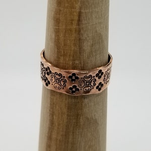 May include: A copper ring with a decorative floral pattern. The ring is on a wooden display stand.