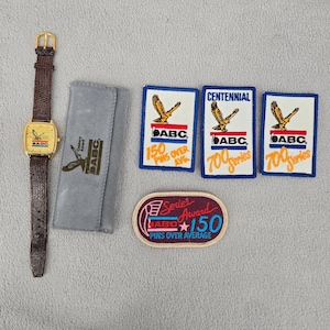 May include: A gold-toned watch with a brown leather band, a gray case, and four patches. The watch face and patches feature the text "ABC" and an eagle emblem. The patches read "150 Pins Over Avg" and "700 Series".