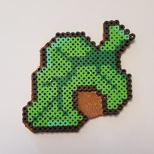 May include: A green and black pixel art design of a leaf, made with small round beads. The design is mounted on a brown background.