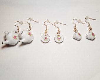May include: Three pairs of gold dangle earrings with porcelain charms. One pair features a teapot, one pair features a saucer, and one pair features a teacup. All charms have a floral design with pink roses.