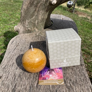 Könnte beinhalten: Eine runde, goldgelbe Kerze mit einem weißen Docht sitzt auf einer Holzoberfläche. Die Kerze befindet sich in einer Schachtel mit einem weißen und grauen geometrischen Muster. Die Schachtel trägt den Text "CANDELA AMICA". Eine kleine Karte mit dem Text "RAHU Medita su questo pianeta e crea spazio al tuo Cosmo Interiore!" befindet sich vor der Kerze.