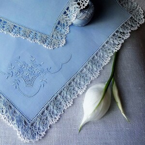 May include: A light blue handkerchief with white crochet trim and an embroidered floral design. The handkerchief is folded and lying on a light blue textured surface.