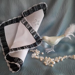 May include: A white handkerchief with black crocheted lace trim. The handkerchief is folded in half and a white porcelain bird with a white feather tail is perched on top of it. A string of white pearls is in the foreground.