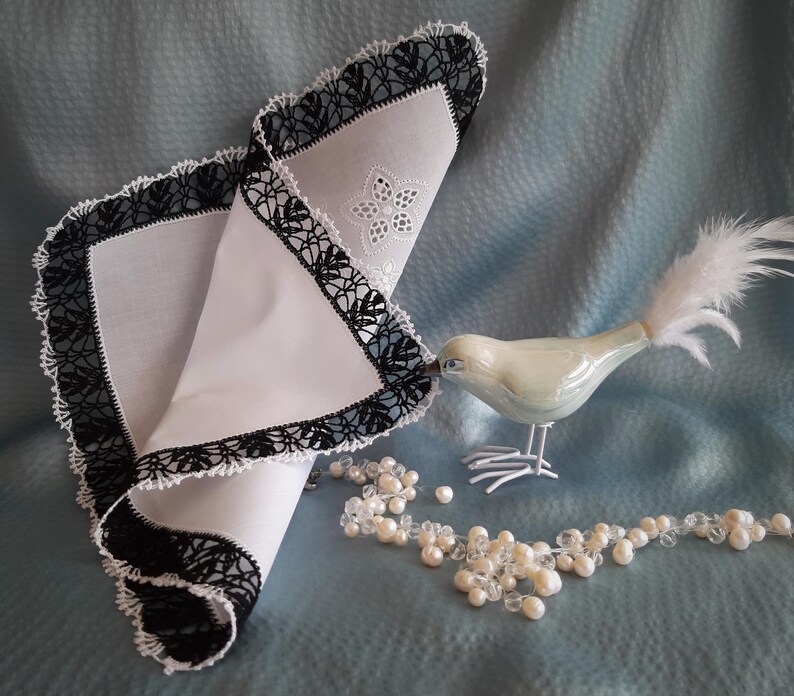 May include: A white handkerchief with black crocheted lace trim. The handkerchief is folded in half and a white porcelain bird with a white feather tail is perched on top of it. A string of white pearls is in the foreground.